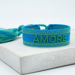 Close-up of adjustable azurro blue woven friendship bracelet with green embroidered "AMORE" lettering, matching green borders, and sliding knot tassel cords.