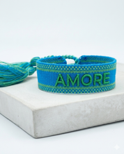 Close-up of adjustable azurro blue woven friendship bracelet with green embroidered "AMORE" lettering, matching green borders, and sliding knot tassel cords.
