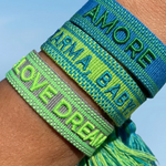 Close-up of adjustable azurro blue woven friendship bracelet with green embroidered "AMORE" lettering, matching green borders, and sliding knot tassel cords.