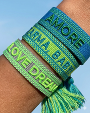 Close-up of adjustable azurro blue woven friendship bracelet with green embroidered "AMORE" lettering, matching green borders, and sliding knot tassel cords.