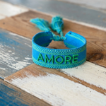 Close-up of adjustable azurro blue woven friendship bracelet with green embroidered "AMORE" lettering, matching green borders, and sliding knot tassel cords.