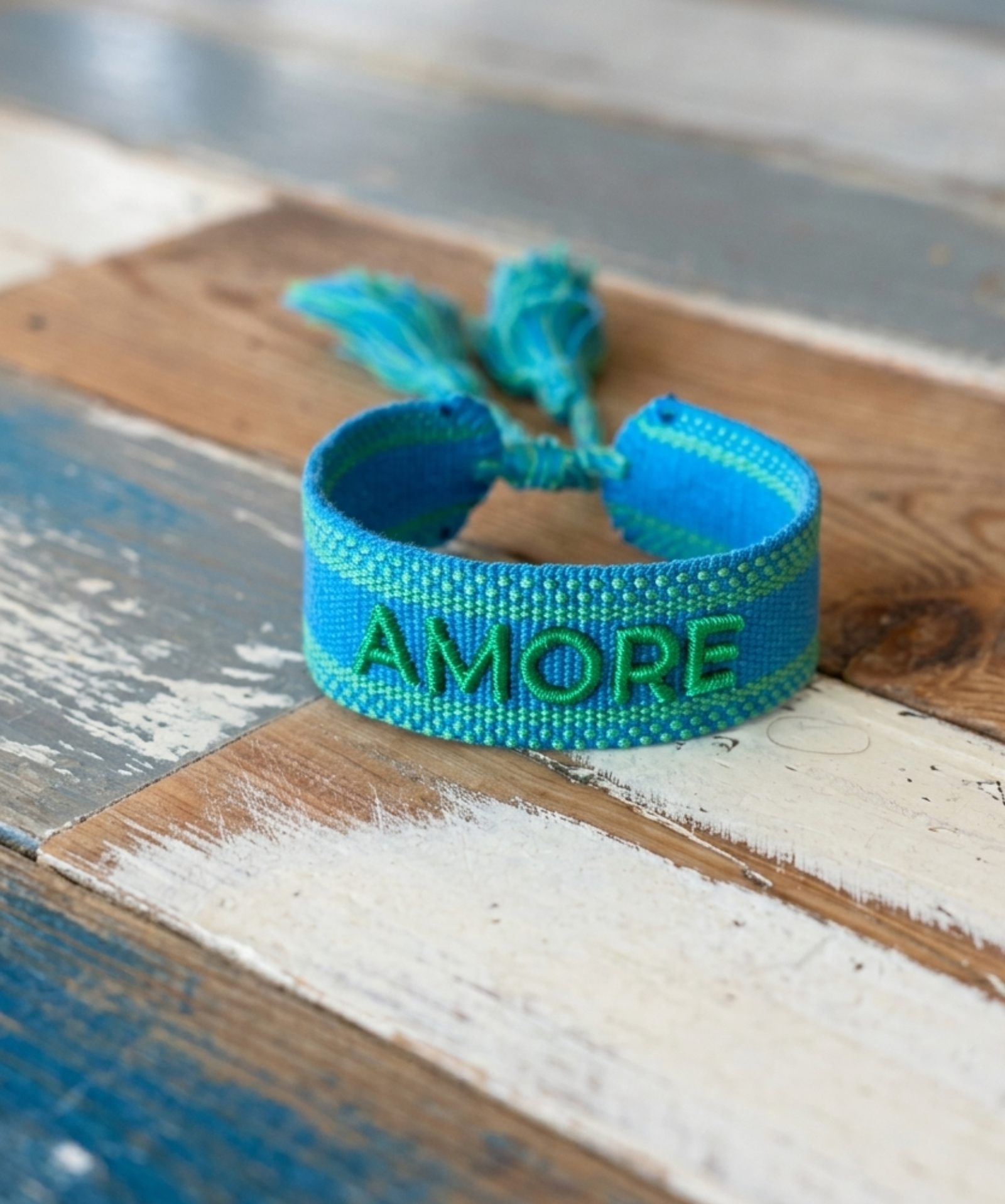 Close-up of adjustable azurro blue woven friendship bracelet with green embroidered "AMORE" lettering, matching green borders, and sliding knot tassel cords.