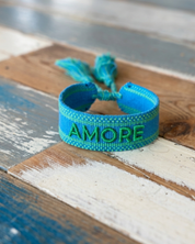 Close-up of adjustable azurro blue woven friendship bracelet with green embroidered "AMORE" lettering, matching green borders, and sliding knot tassel cords.