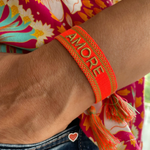 Close-up of adjustable neon orange woven friendship bracelet with green border and gold embroidered "AMORE" lettering and sliding knot cords.