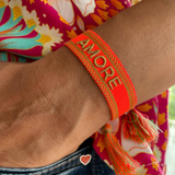 Close-up of adjustable neon orange woven friendship bracelet with green border and gold embroidered "AMORE" lettering and sliding knot cords.