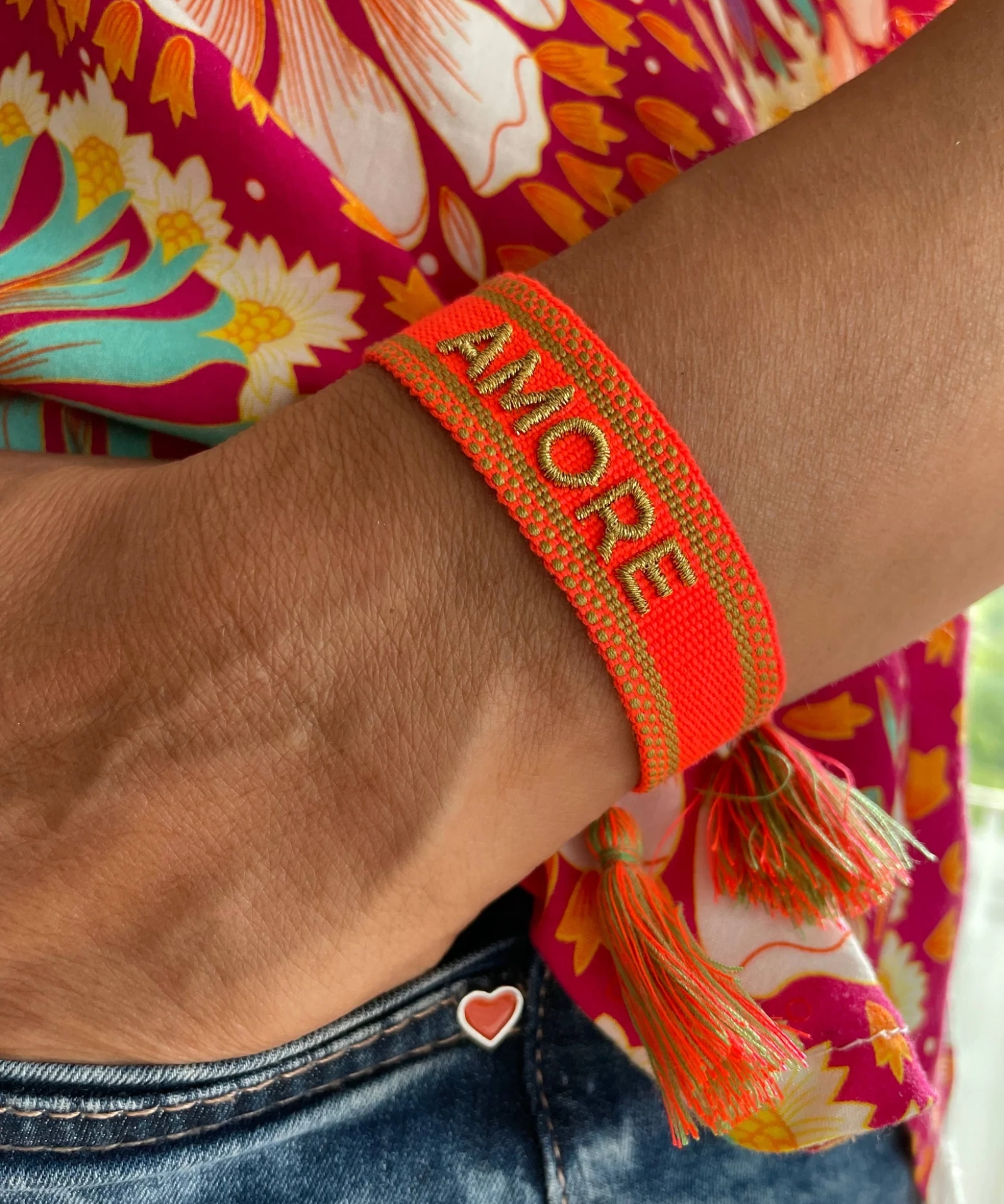 Close-up of adjustable neon orange woven friendship bracelet with green border and gold embroidered "AMORE" lettering and sliding knot cords.