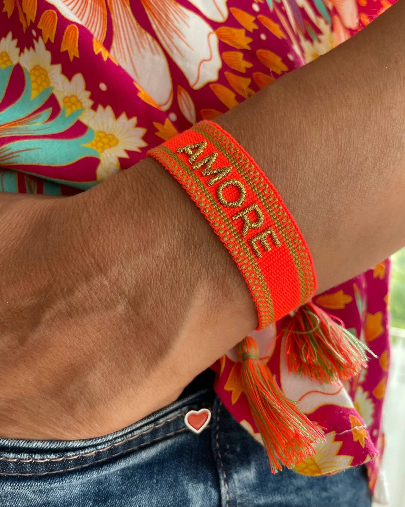 Close-up of adjustable neon orange woven friendship bracelet with green border and gold embroidered "AMORE" lettering and sliding knot cords.