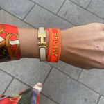 Close-up of adjustable neon orange woven friendship bracelet with green border and gold embroidered "AMORE" lettering and sliding knot cords.