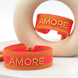 Close-up of adjustable neon orange woven friendship bracelet with green border and gold embroidered "AMORE" lettering and sliding knot cords.