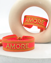 Close-up of adjustable neon orange woven friendship bracelet with green border and gold embroidered "AMORE" lettering and sliding knot cords.