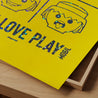 A close-up of an iconic figurine portrait on an official Playmobil-licensed art print with the slogan Live - Love - Play Mobil in metallic hot print on vibrant yellow paper.