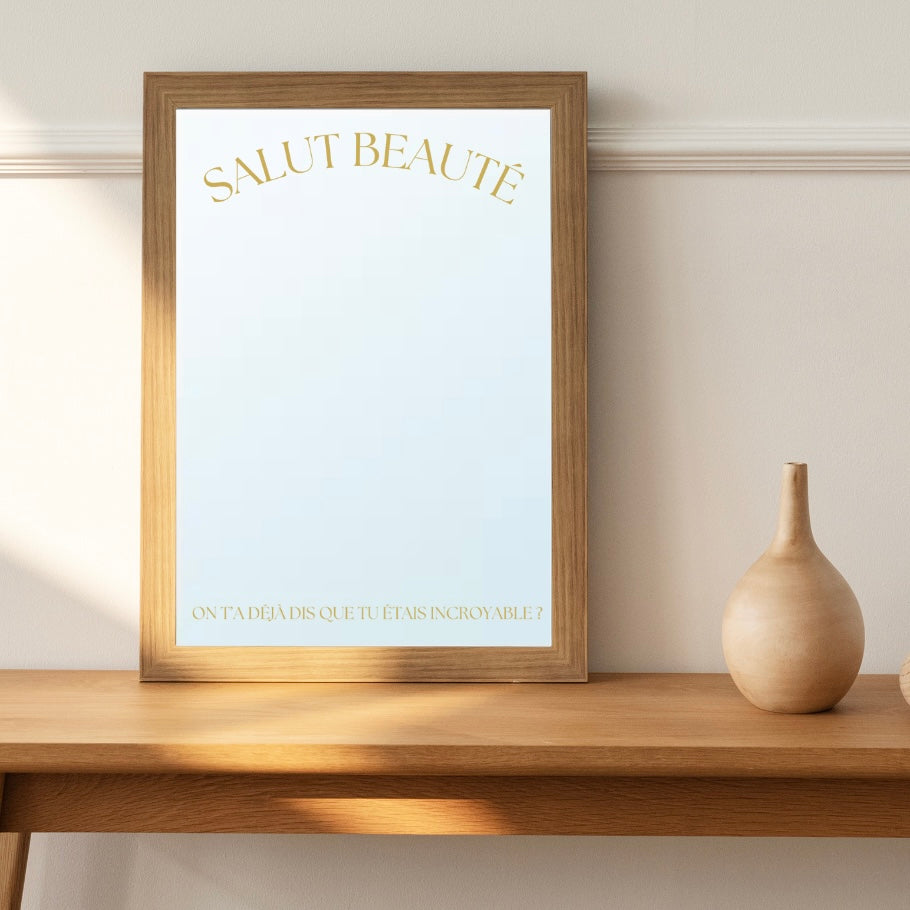 A mirror by the French brand Atelier Circe, adorned with a message reading 'Hello Beauty' in gold letters, is situated on a table adjacent to a vase. 