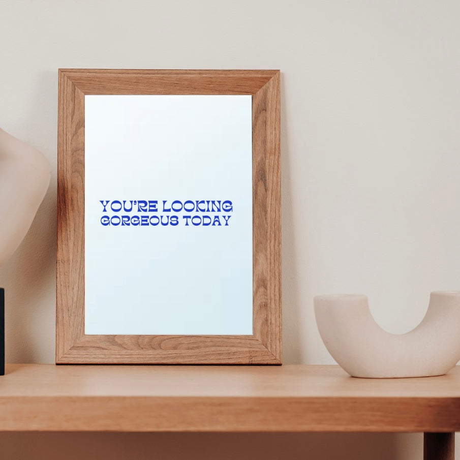 A mirror by the French brand Atelier Circe, adorned with a message reading 'Gorgeous' in blue lettering, on a table adjacent to a vase. 