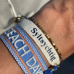 Close-up of adjustable blue woven friendship bracelet with off-white and orange border and embroidered "BEACH DAYS" lettering with sliding knot cords.