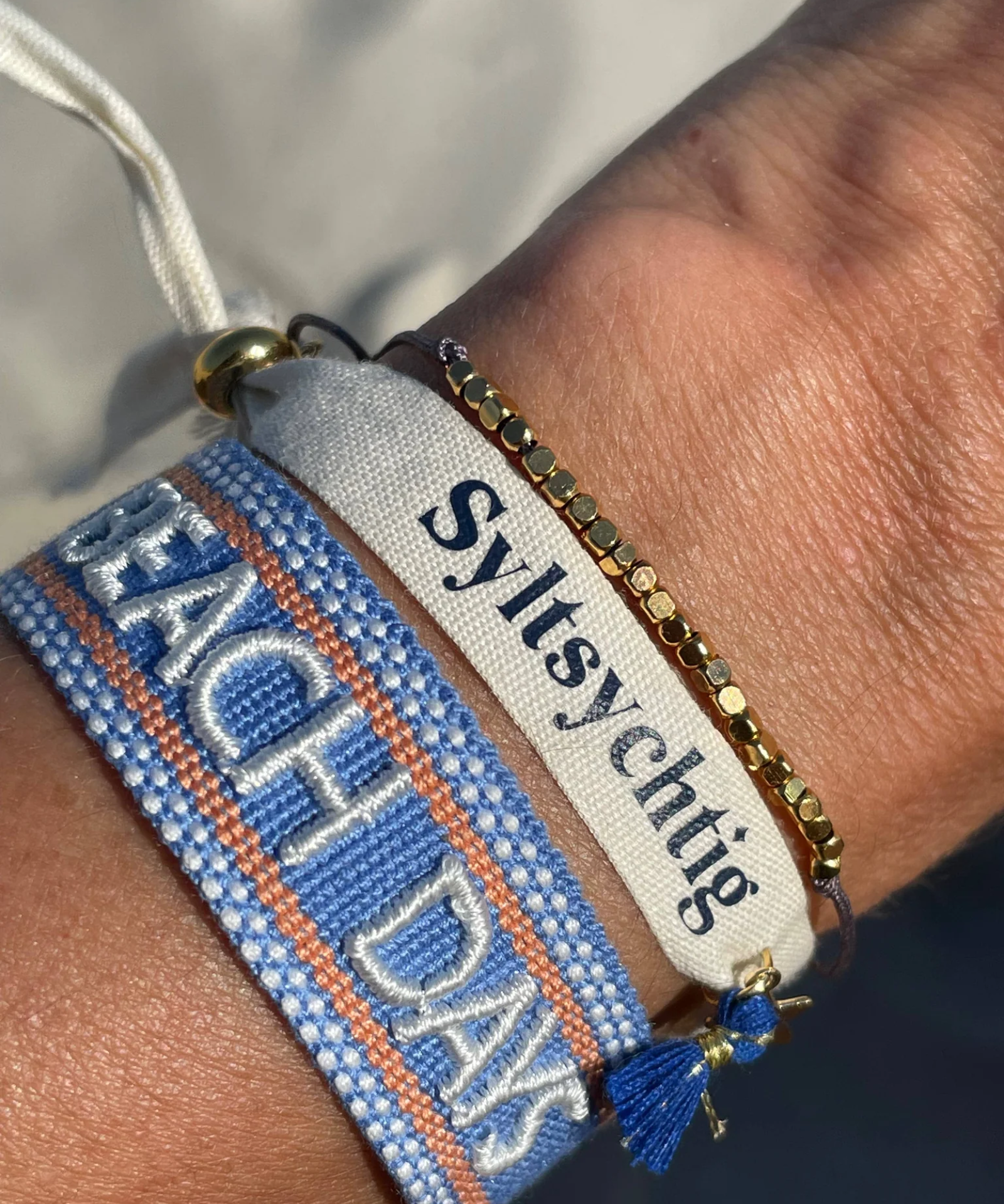 Close-up of adjustable blue woven friendship bracelet with off-white and orange border and embroidered "BEACH DAYS" lettering with sliding knot cords.