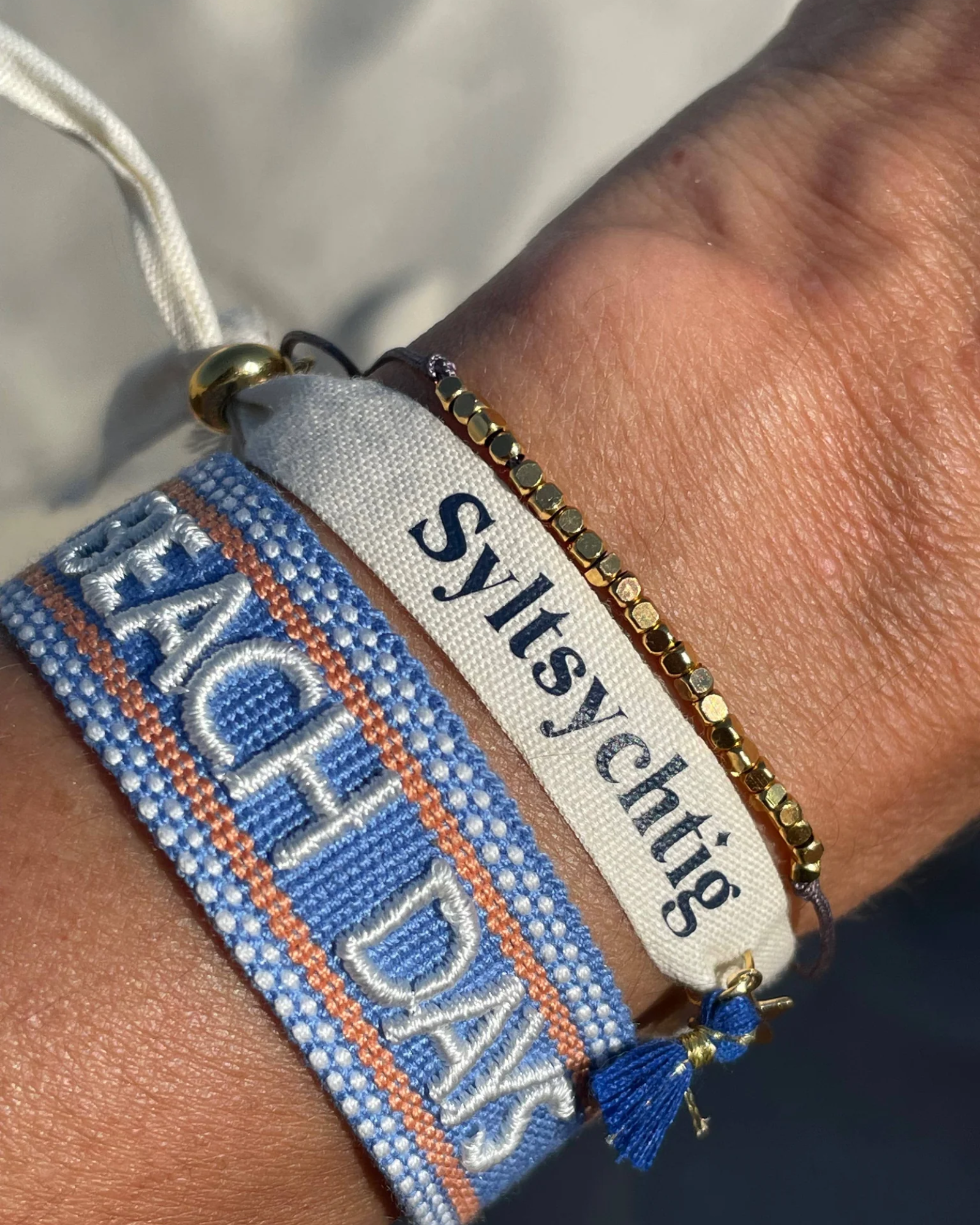 Close-up of adjustable blue woven friendship bracelet with off-white and orange border and embroidered "BEACH DAYS" lettering with sliding knot cords.