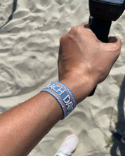 Close-up of adjustable blue woven friendship bracelet with off-white and orange border and embroidered "BEACH DAYS" lettering with sliding knot cords.