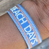 Close-up of adjustable blue woven friendship bracelet with off-white and orange border and embroidered "BEACH DAYS" lettering with sliding knot cords.