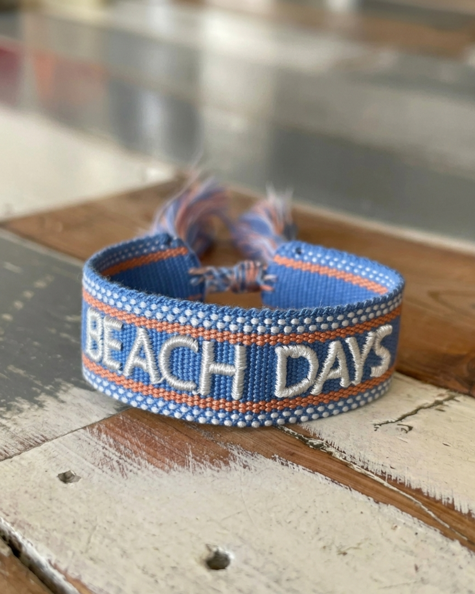 Close-up of adjustable blue woven friendship bracelet with off-white and orange border and embroidered "BEACH DAYS" lettering with sliding knot cords.