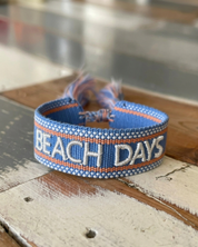 Close-up of adjustable blue woven friendship bracelet with off-white and orange border and embroidered "BEACH DAYS" lettering with sliding knot cords.
