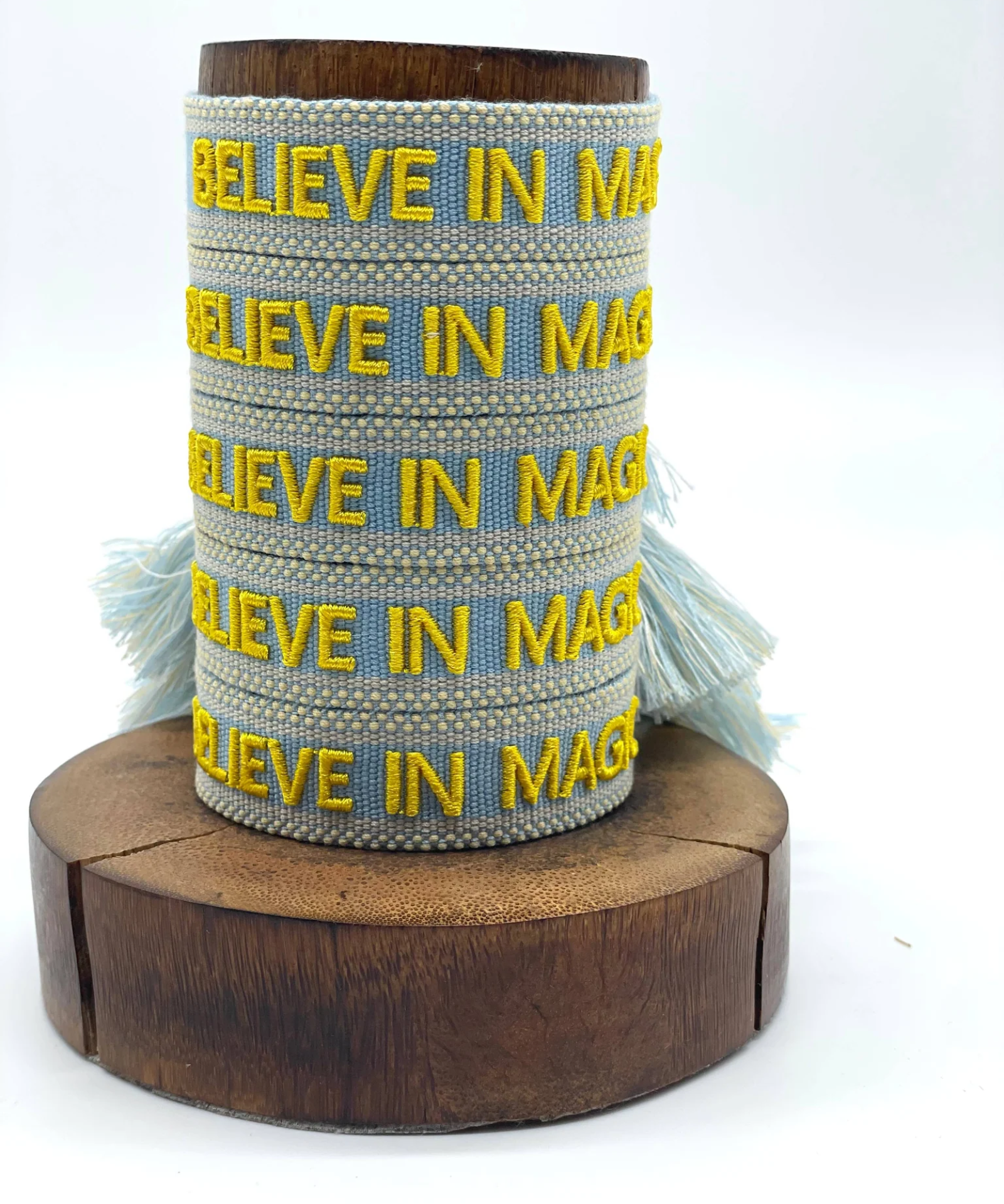 Close-up of adjustable light blue woven friendship bracelet with yellow embroidered "BELIEVE IN MAGIC" text and off-white border with sliding knot cords.