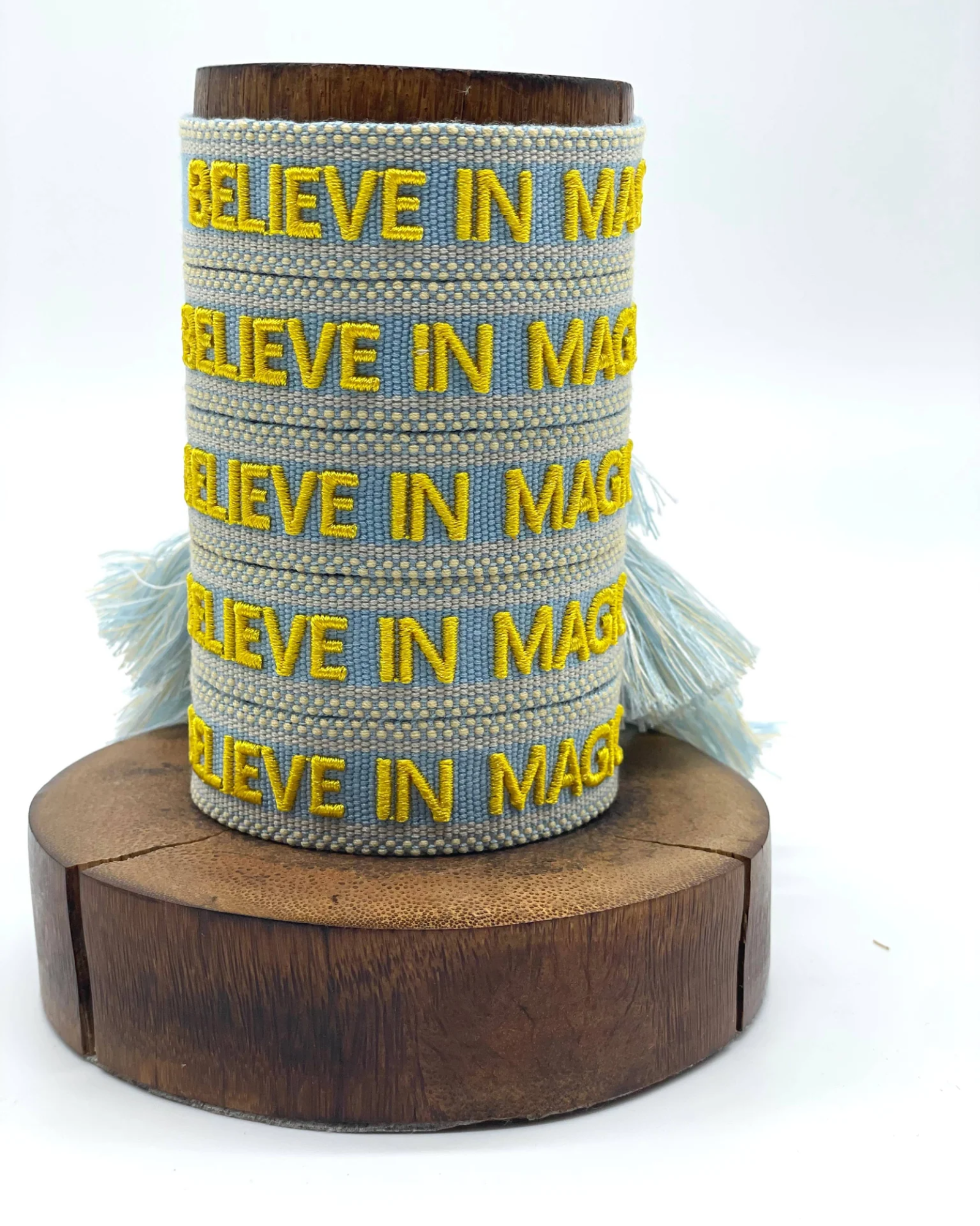 Close-up of adjustable light blue woven friendship bracelet with yellow embroidered "BELIEVE IN MAGIC" text and off-white border with sliding knot cords.