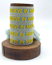 Close-up of adjustable light blue woven friendship bracelet with yellow embroidered "BELIEVE IN MAGIC" text and off-white border with sliding knot cords.