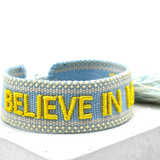 Close-up of adjustable light blue woven friendship bracelet with yellow embroidered "BELIEVE IN MAGIC" text and off-white border with sliding knot cords.