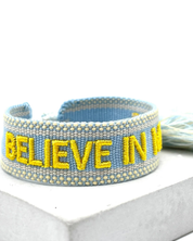 Close-up of adjustable light blue woven friendship bracelet with yellow embroidered "BELIEVE IN MAGIC" text and off-white border with sliding knot cords.
