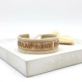 Close-up of adjustable beige woven bracelet with glitter taupe embroidered “CHAMPAGNE BABY” lettering, glitter taupe gold border, and sliding knot tassel cords.