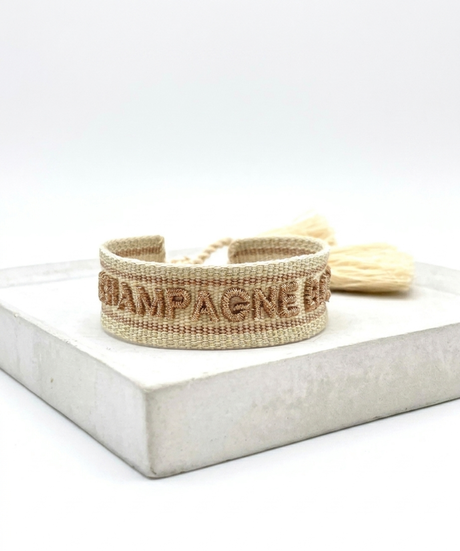 Close-up of adjustable beige woven bracelet with glitter taupe embroidered “CHAMPAGNE BABY” lettering, glitter taupe gold border, and sliding knot tassel cords.