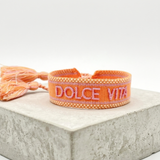 Close-up of adjustable orange woven bracelet with embroidered “DOLCE VITA” lettering and sliding knot tassel cords.