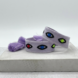 Close-up of adjustable lavender woven bracelet featuring embroidered evil eye talisman design with sliding knot tassel cords.