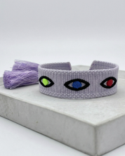 Close-up of adjustable lavender woven bracelet featuring embroidered evil eye talisman design with sliding knot tassel cords.
