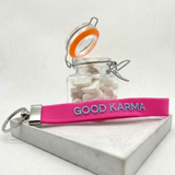 Close-up of embroidered pink woven keychain with blue “GOOD KARMA” text and silver metal keyring on a tightly woven strap.