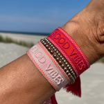Close-up of adjustable rosy peach woven bracelet with embroidered “GOOD VIBES” lettering and sliding knot tassel cords.