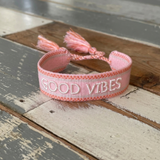 Close-up of adjustable rosy peach woven bracelet with embroidered “GOOD VIBES” lettering and sliding knot tassel cords.