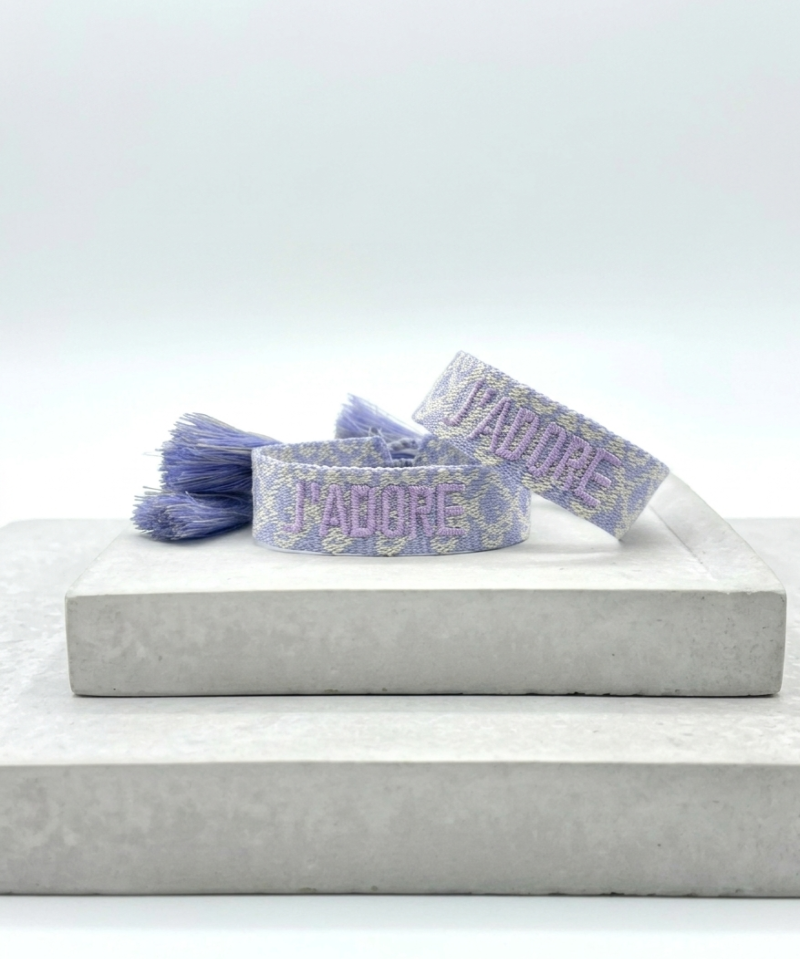 Close-up of adjustable purple and cream jacquard woven friendship bracelet with lilac embroidered “J’ADORE” lettering and sliding knot tassel cords.