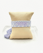 Close-up of adjustable purple and cream jacquard woven friendship bracelet with lilac embroidered “J’ADORE” lettering and sliding knot tassel cords.