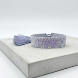 Close-up of adjustable lilac jacquard woven bracelet with embroidered “J'ADORE” lettering and sliding knot tassel cords.