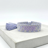 Close-up of adjustable purple and cream jacquard woven friendship bracelet with lilac embroidered “J’ADORE” lettering and sliding knot tassel cords.