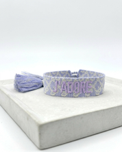 Close-up of adjustable purple and cream jacquard woven friendship bracelet with lilac embroidered “J’ADORE” lettering and sliding knot tassel cords.
