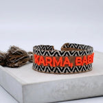 Close-up of adjustable woven boho friendship bracelet with neon orange “Karma, Baby!” embroidery and sliding tassel cords on a brown woven band.