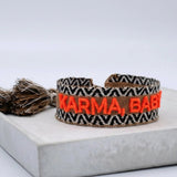 Close-up of adjustable woven boho friendship bracelet with neon orange “Karma, Baby!” embroidery and sliding tassel cords on a brown woven band.