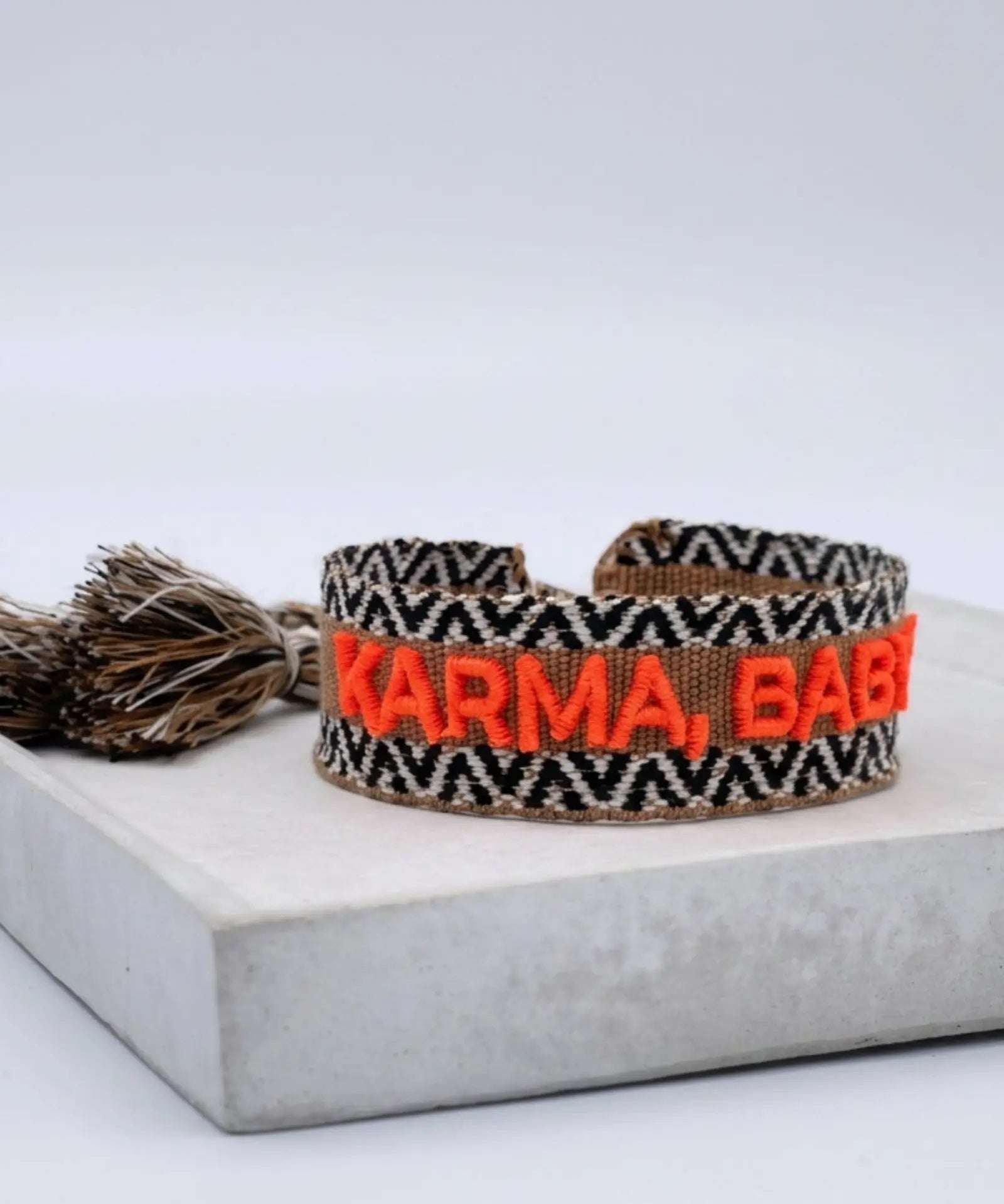 Close-up of adjustable woven boho friendship bracelet with neon orange “Karma, Baby!” embroidery and sliding tassel cords on a brown woven band.