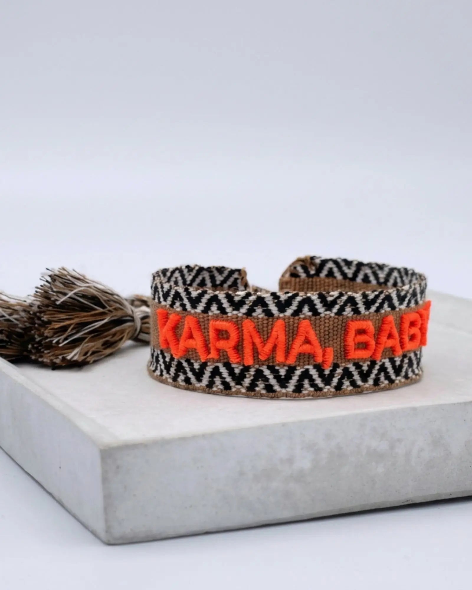 Close-up of adjustable woven boho friendship bracelet with neon orange “Karma, Baby!” embroidery and sliding tassel cords on a brown woven band.
