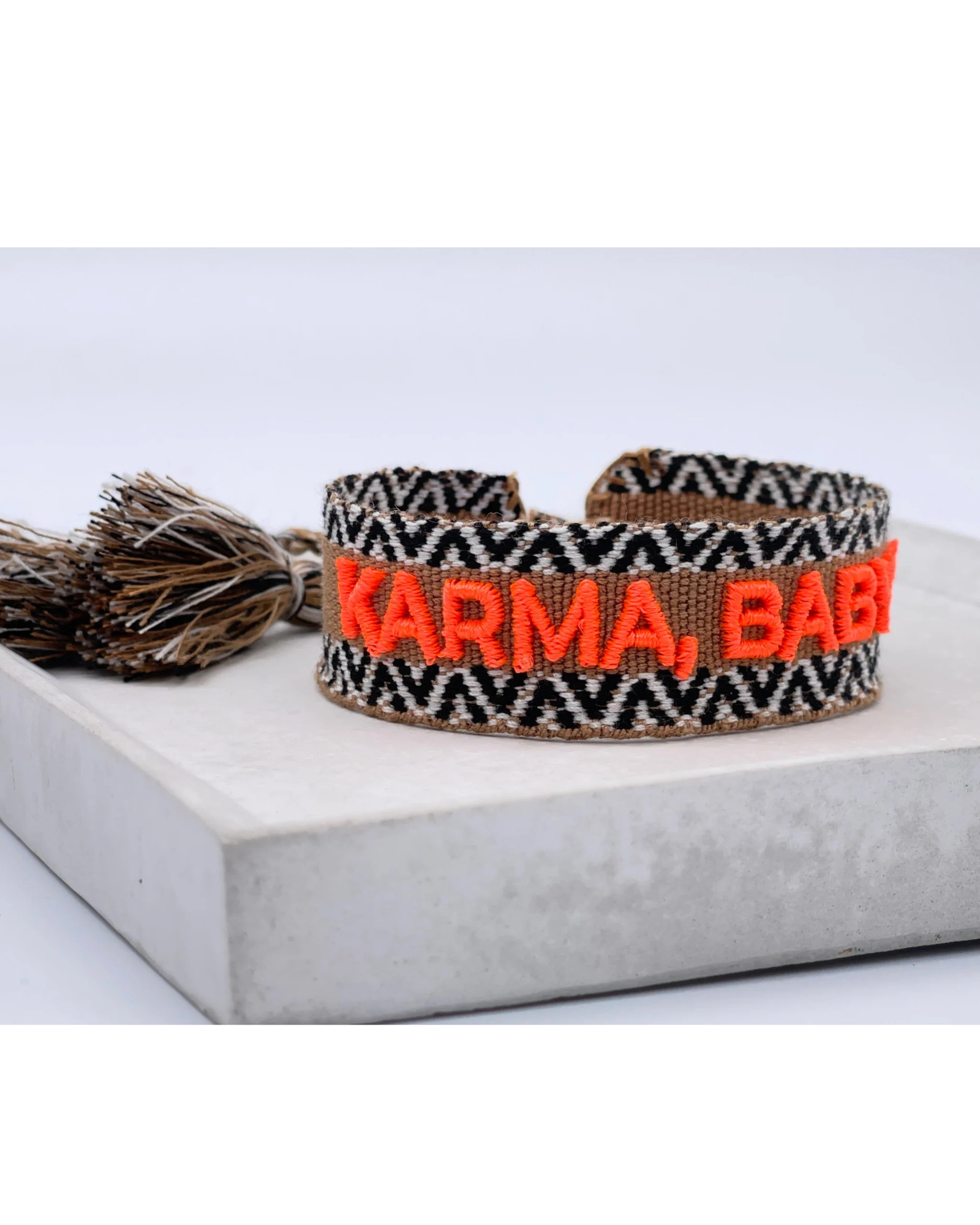 Close-up of adjustable woven boho friendship bracelet with neon orange “Karma, Baby!” embroidery and sliding tassel cords on a brown woven band.