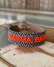 Close-up of adjustable woven boho friendship bracelet with neon orange “Karma, Baby!” embroidery and sliding tassel cords on a brown woven band.