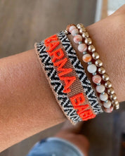 Close-up of adjustable woven boho friendship bracelet with neon orange “Karma, Baby!” embroidery and sliding tassel cords on a brown woven band.