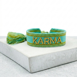 Close-up of adjustable turquoise woven bracelet with gold embroidered “KARMA” lettering and sliding knot tassel cords.